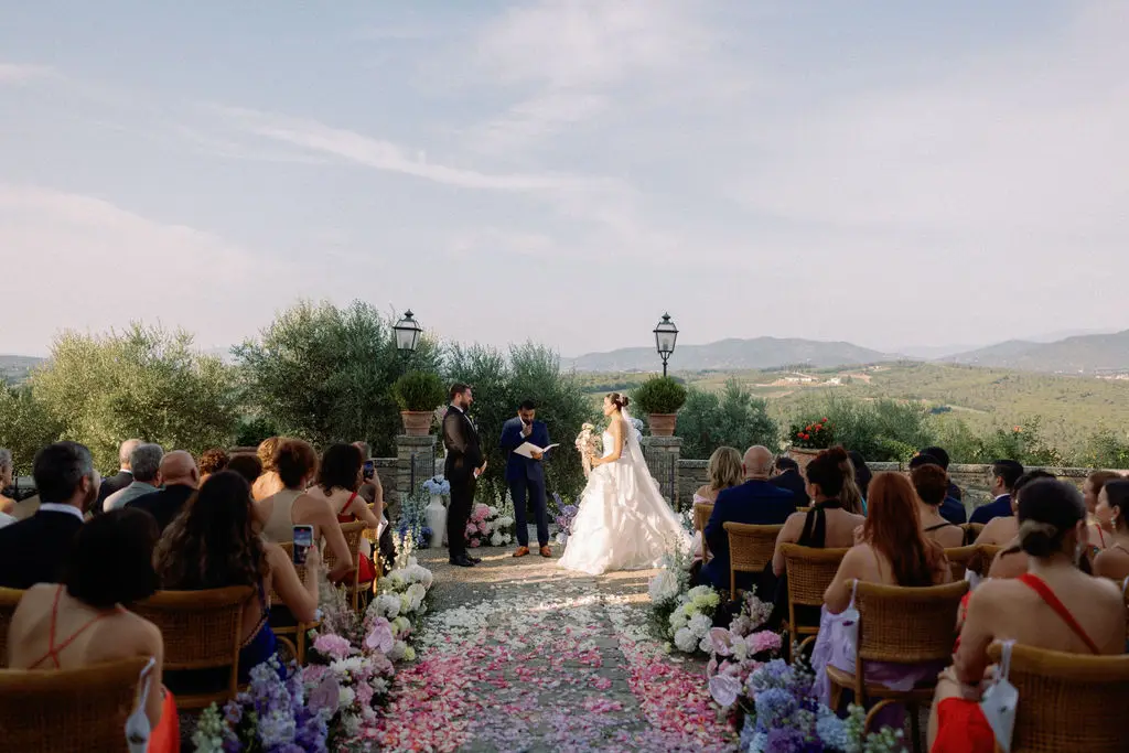 A picturesque garden wedding during a summer late afternoon, with a bride and groom standing at the center surrounded by pastel-toned flowers lining the ground. Guests are seated, facing the couple, as flower petals scatter across the aisle. The backdrop features serene fields and rolling hills under the warm, golden light of the setting sun, creating a romantic and whimsical atmosphere.